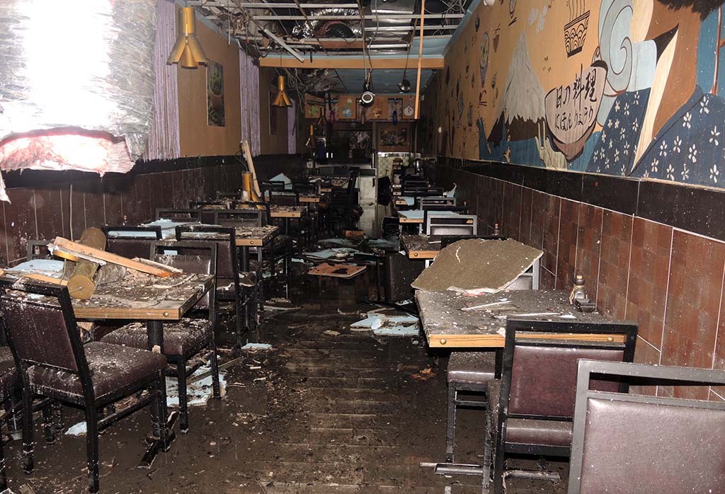 Chairs and tables with pieces of ceiling on them, various debris in the narrow dining area of restaurant